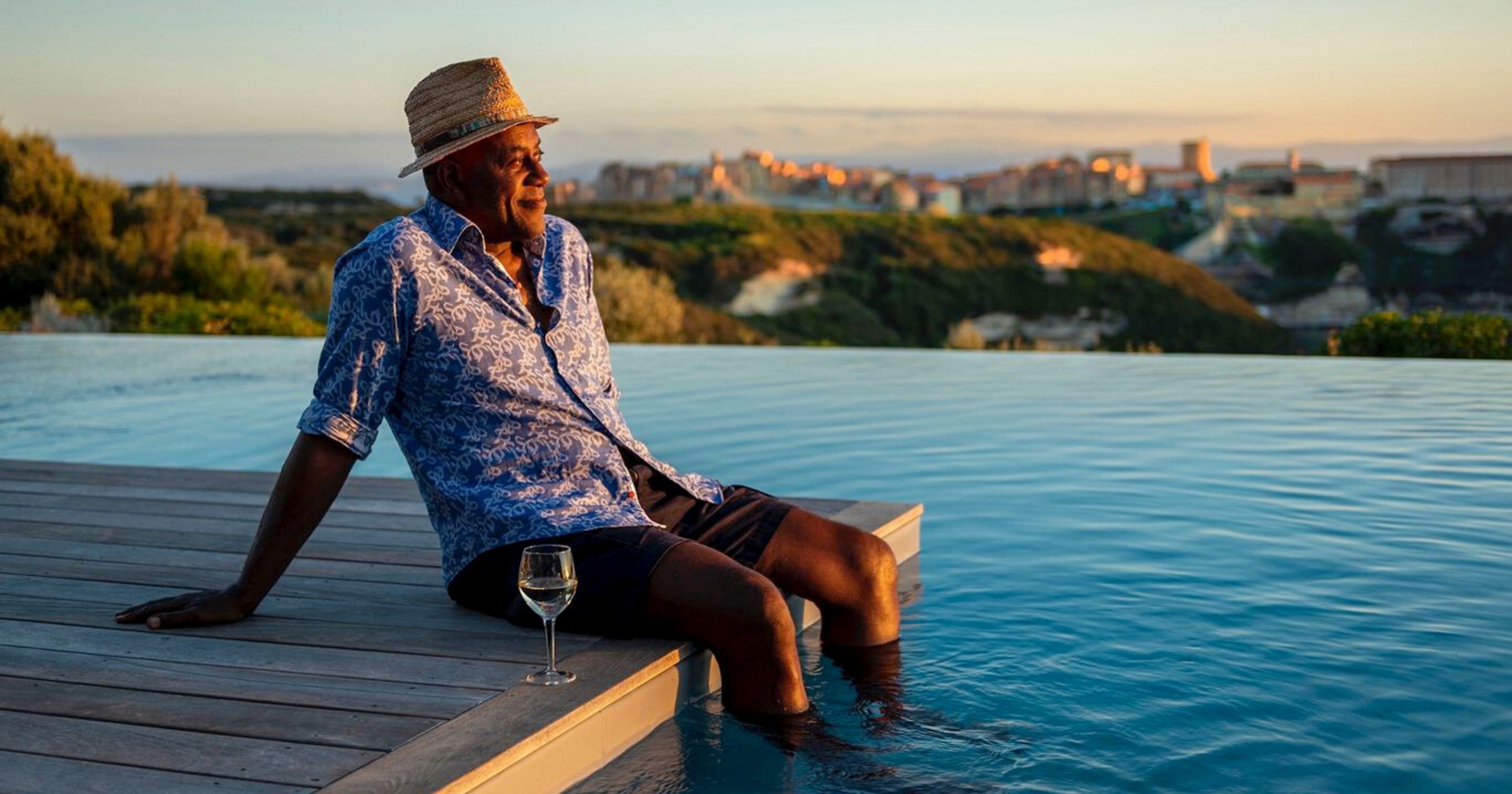 Ainsley Harriot sits with his legs in a swimming pool enjoying a glass of wine