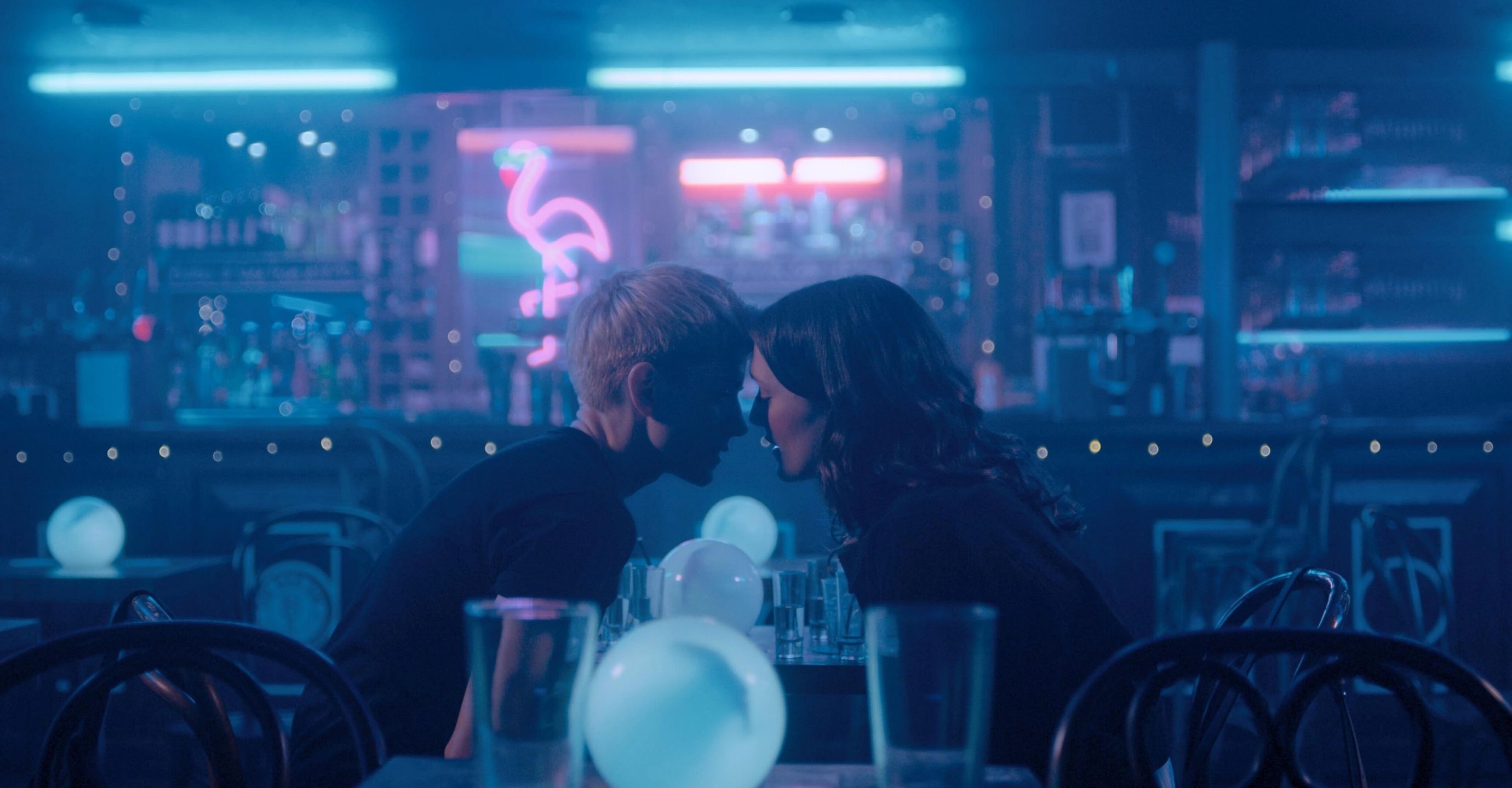 Two woman sit in an empty bar leaning in before a kiss