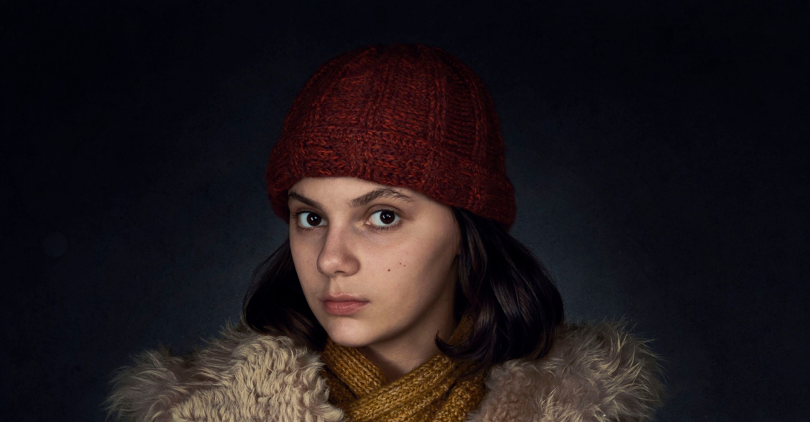 Young Lyra dressed in a heavy fur coat and bobble hat on a dark background