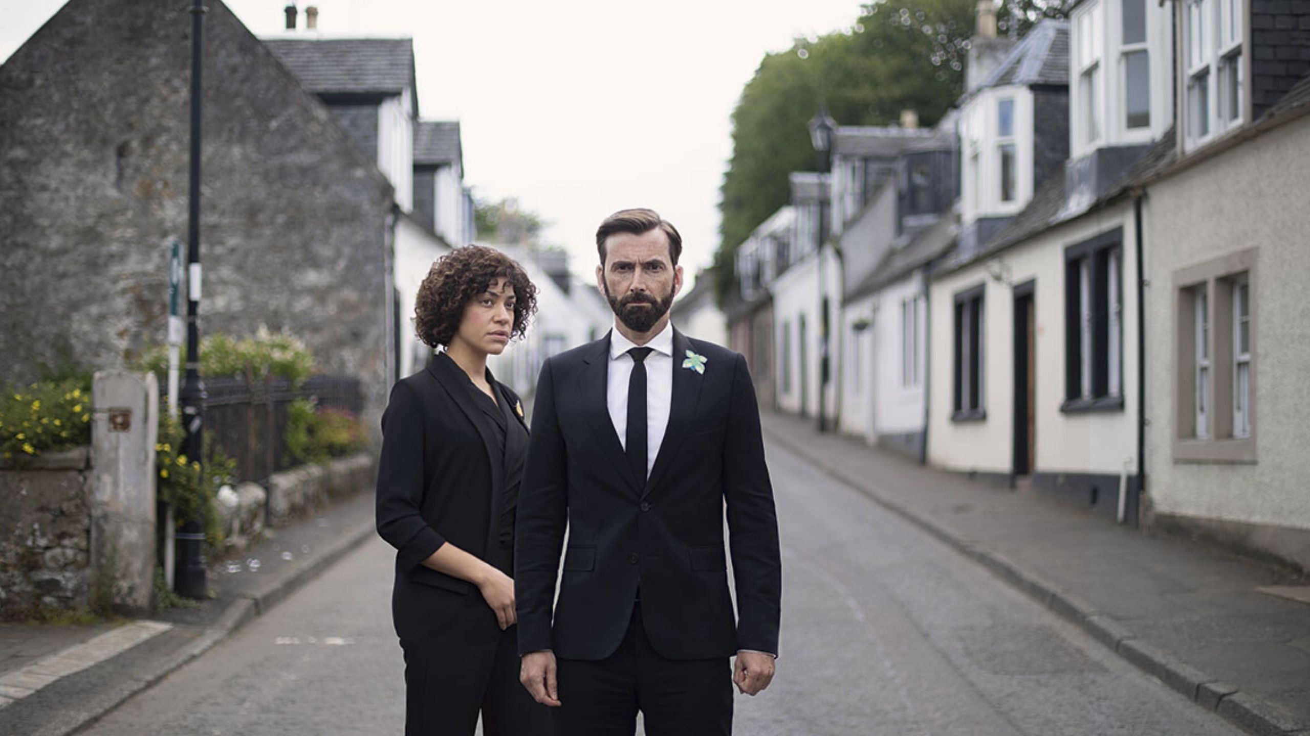 David Tenant stands in the middle of a street looking at the camera, a woman stands at his side looking towards him