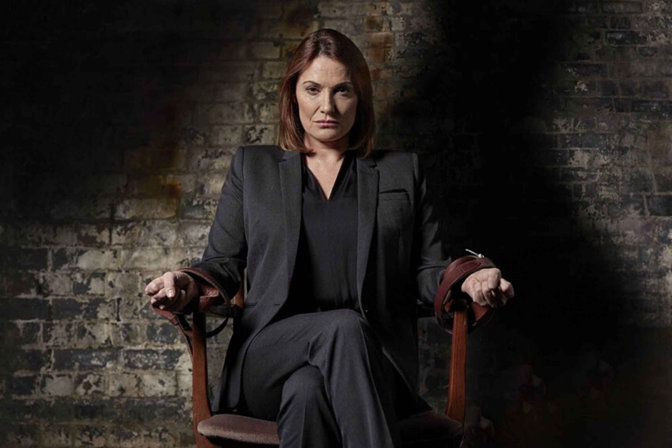Woman in a suit tied to a chair, against a dark brick background