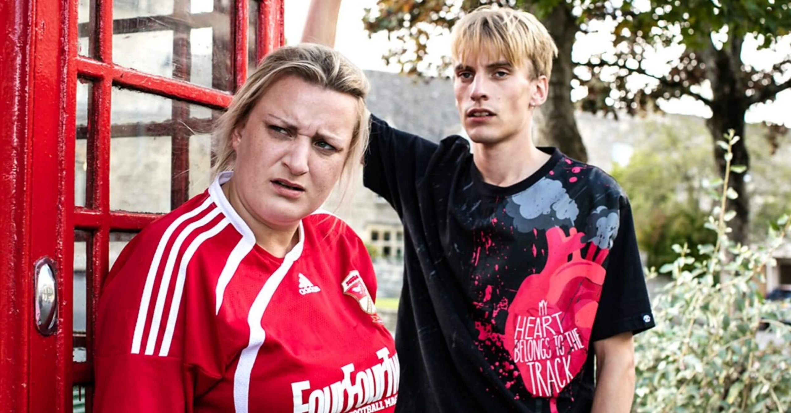 Two young adults stand near a phone box surrounded by countryside