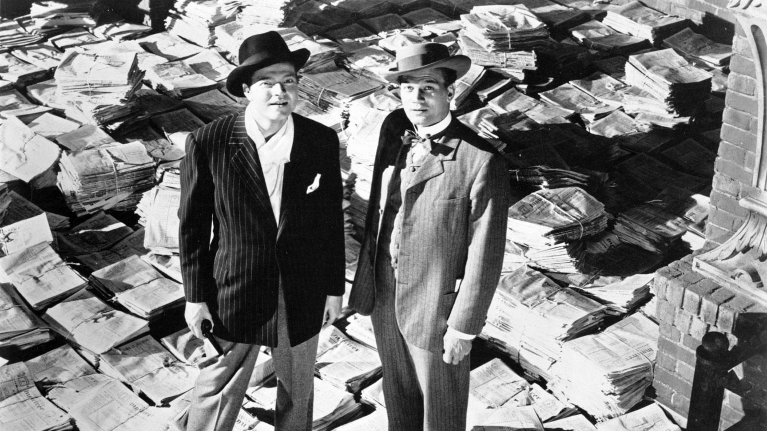 Two men stand on a floor strewn with newspapers looking up at the ceiling