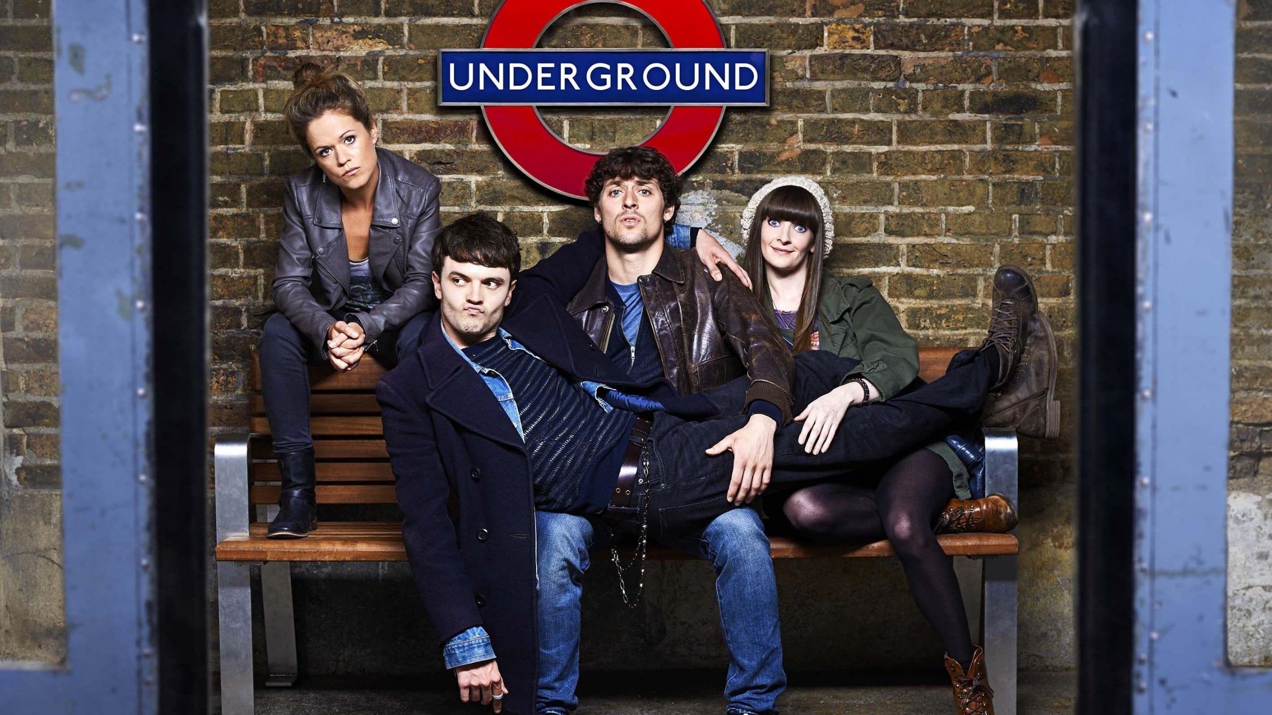 Group of young adults sit looking fed up on a bench in London Underground, one man lays across the legs of the rest of the group