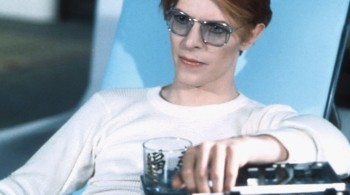 David Bowie starring in The Man who Fell to the Earth, sat on chair wearing blue tinted sunglasses holding a drink