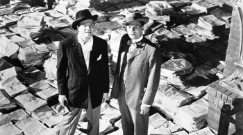 Two men stand on a floor strewn with newspapers looking up at the ceiling