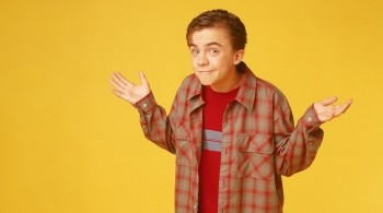 A young Caucasian boy stands against a yellow background holding his hands up in the air looking confused