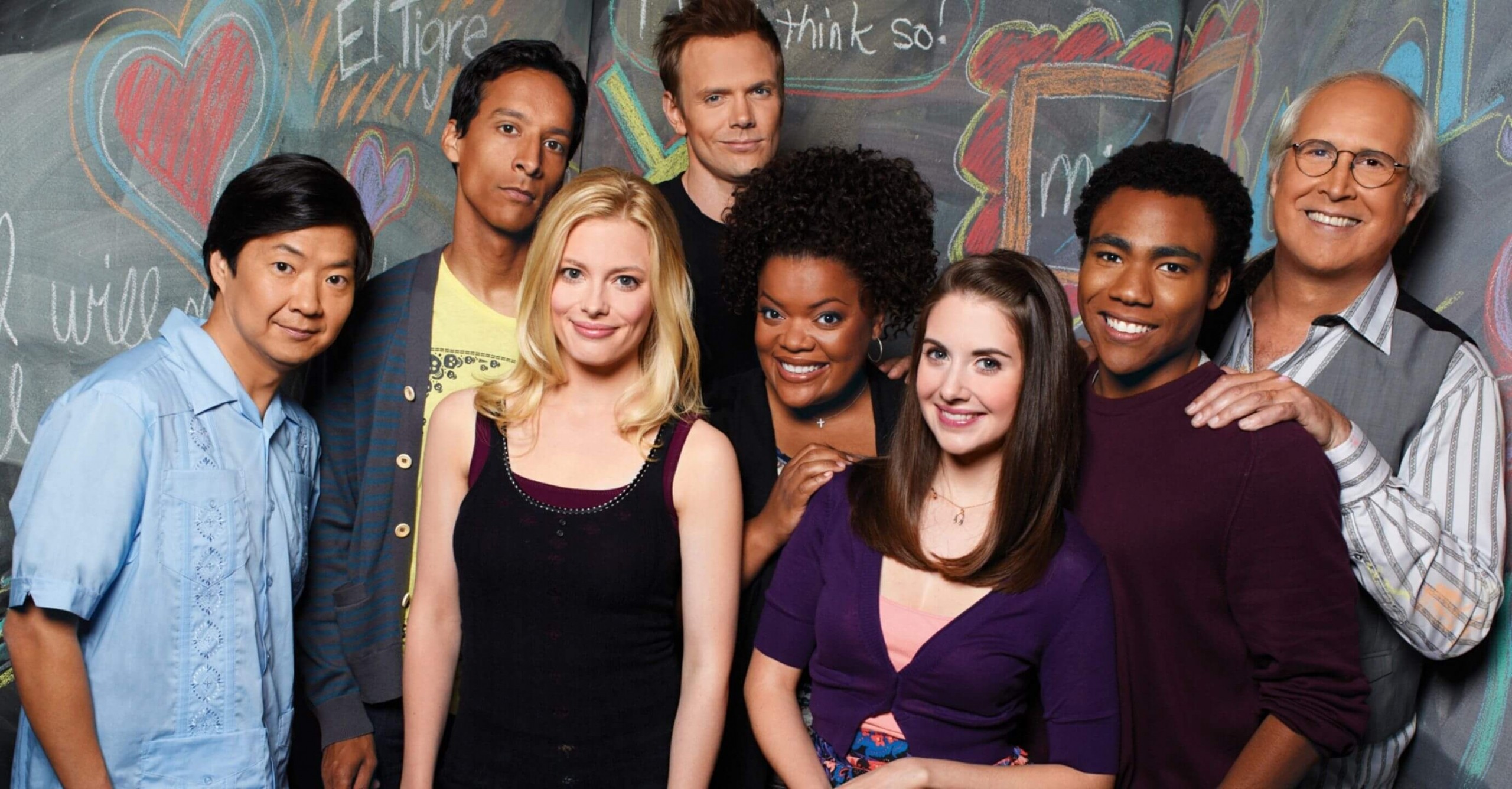 The cast of Community stood in front of a chalk board covered in doodles