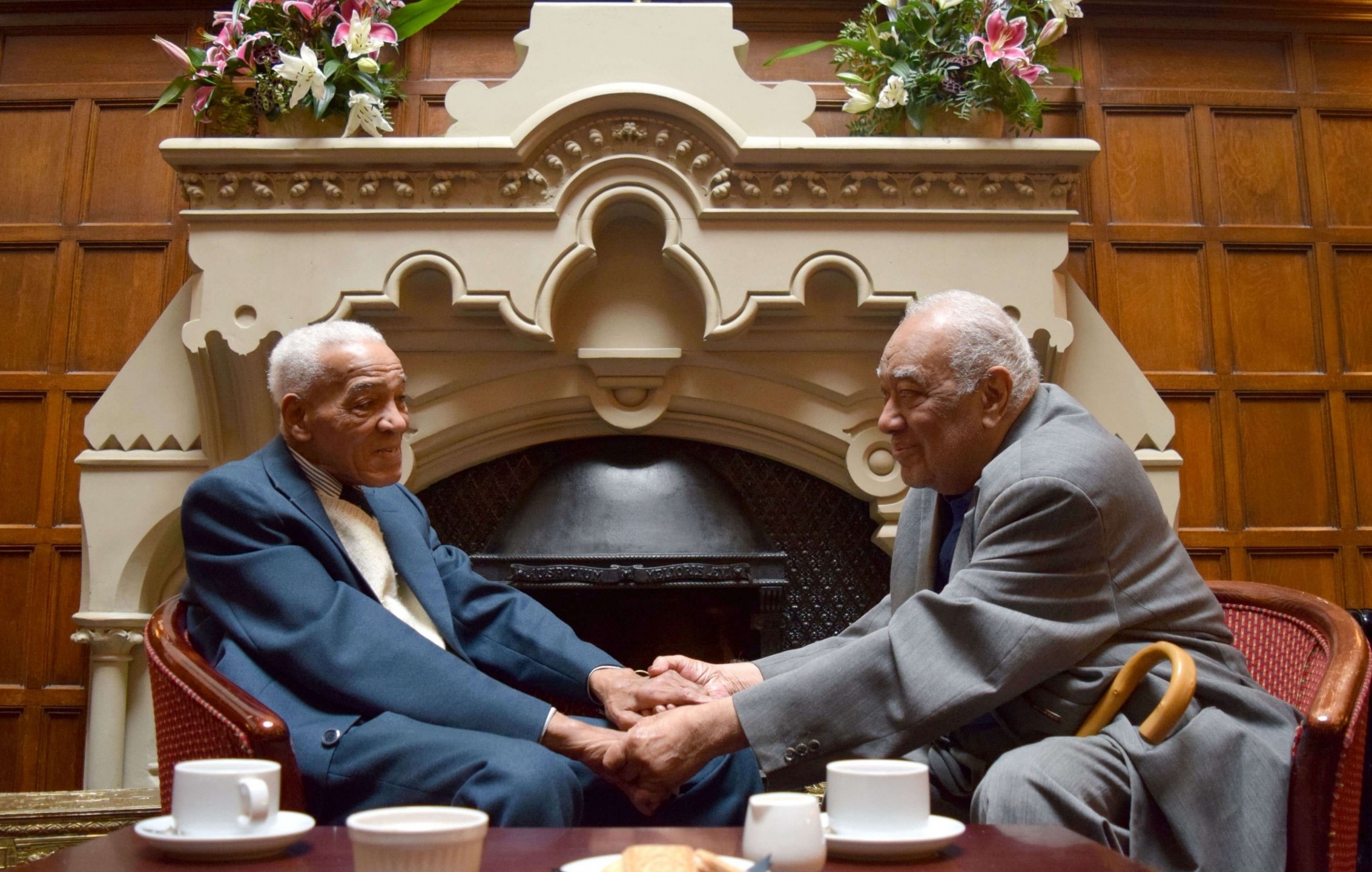 Two elderly Caribbean men, sat down and holding hands in front of a fire place