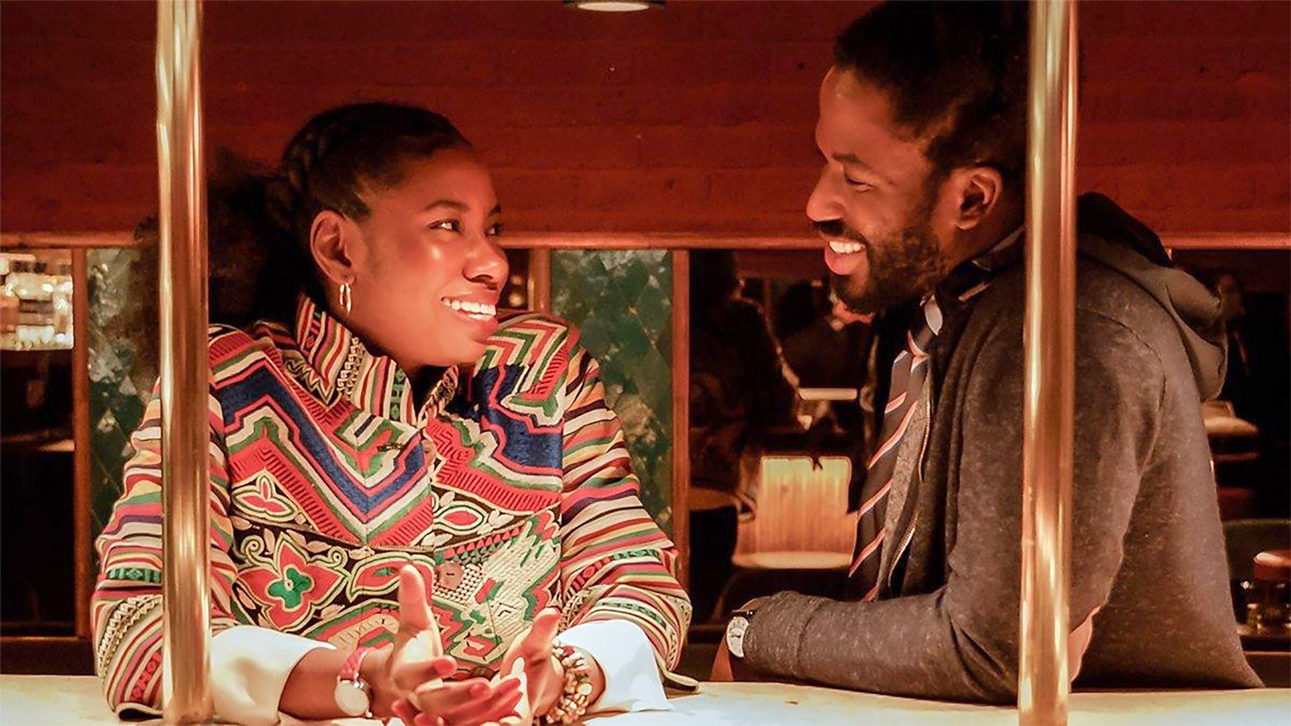 A young Black couple grin at each other behind a bar