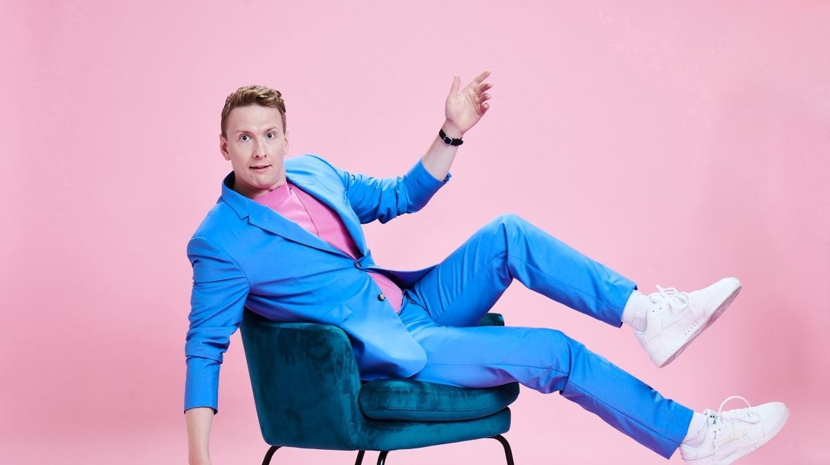 Joe Lycett in a bright blue suit sat jauntily on a chair against a bright pink background