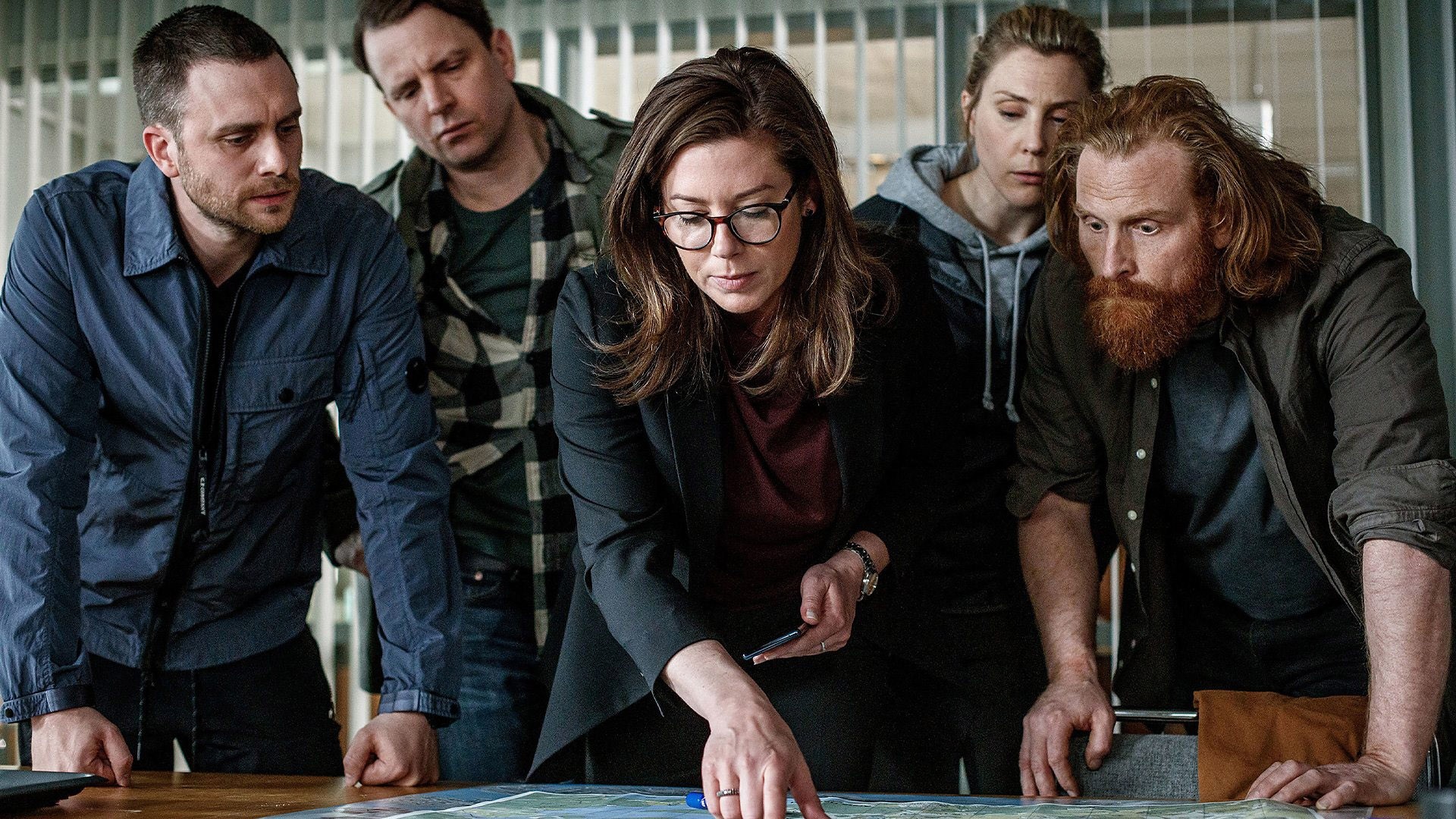Five detectives gathered around a table discussing evidence
