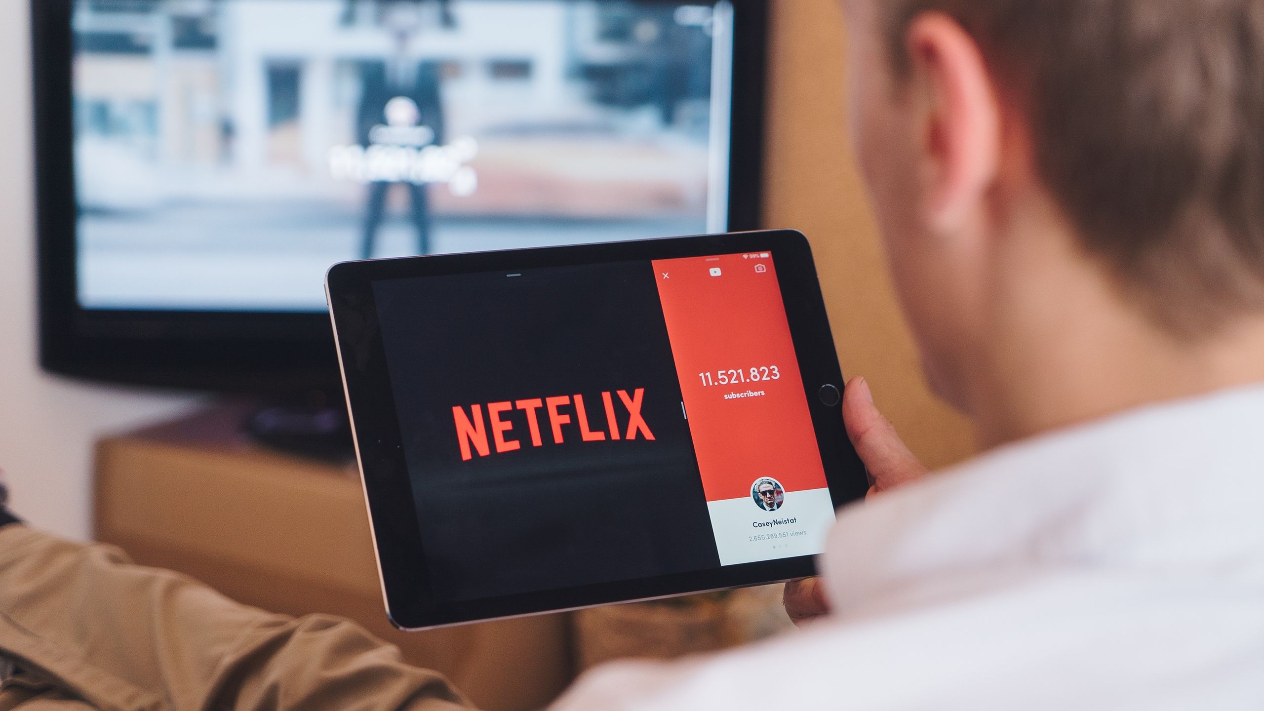 Image of a man streaming Netflix via his tablet with his TV on in the background
